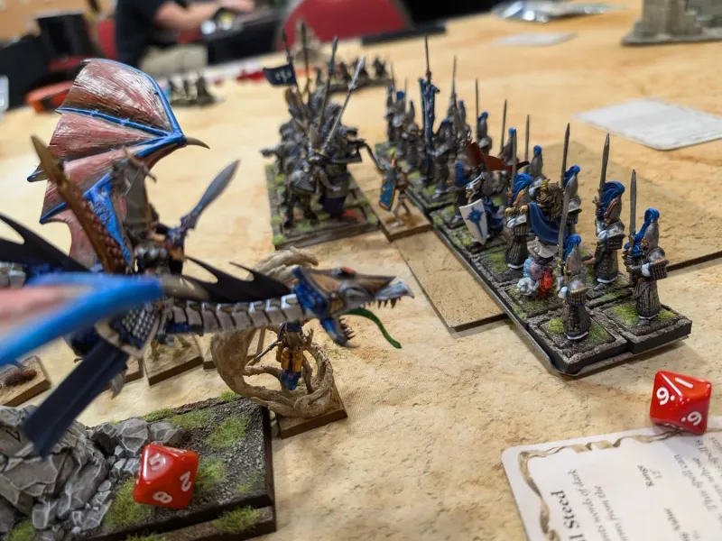 A blue Tomb Kings bone dragon facing ranked High Elf spearmen and a chariot across a sandy tournament table.