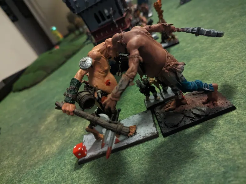 Two giant miniatures locked in combat on a green gaming mat, one swinging a bundled-log club at the other.