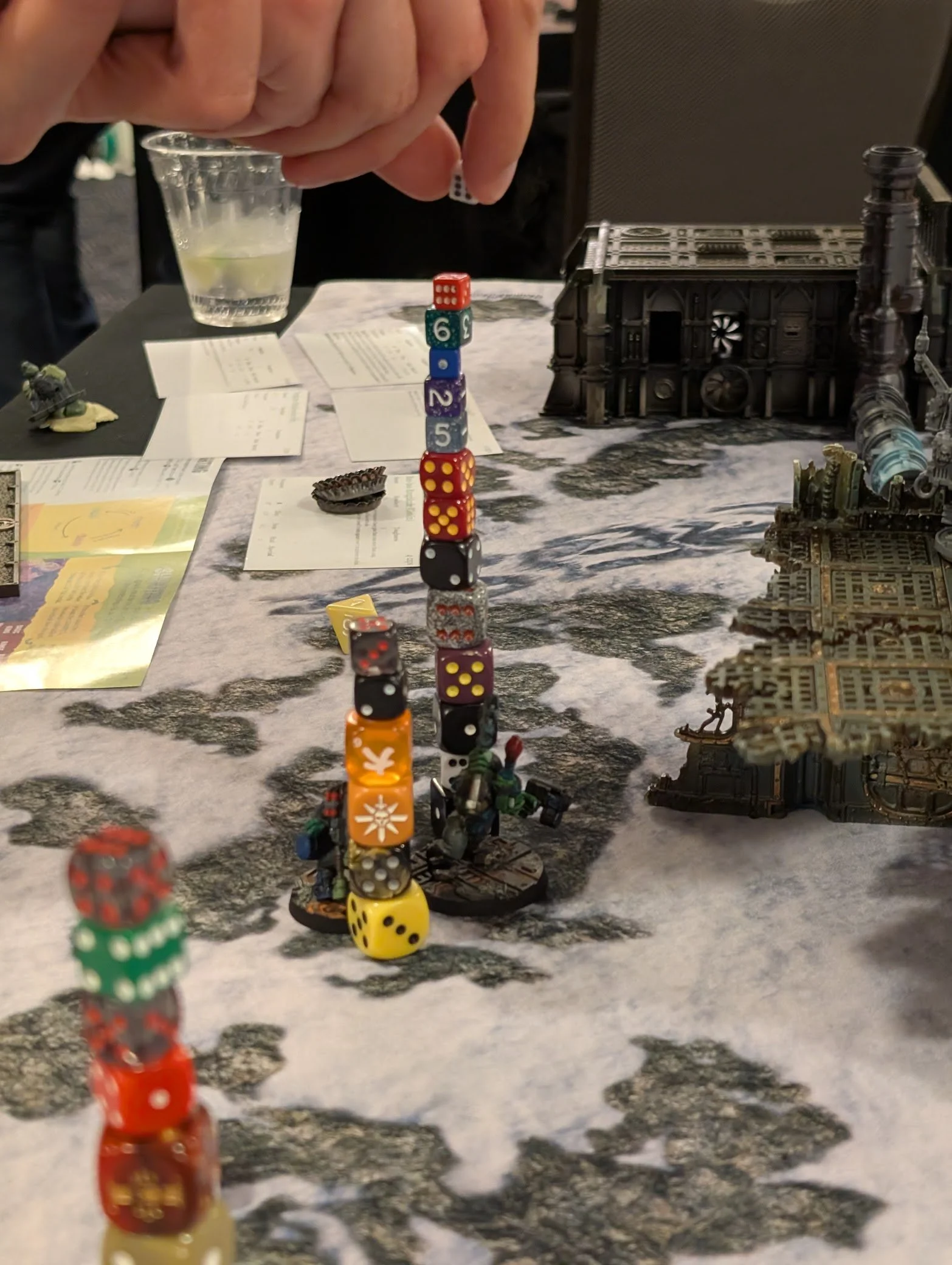 A hand balancing another die on top of a tall stack of mismatched dice rising from an ork miniature, with gothic terrain on the convention table behind.