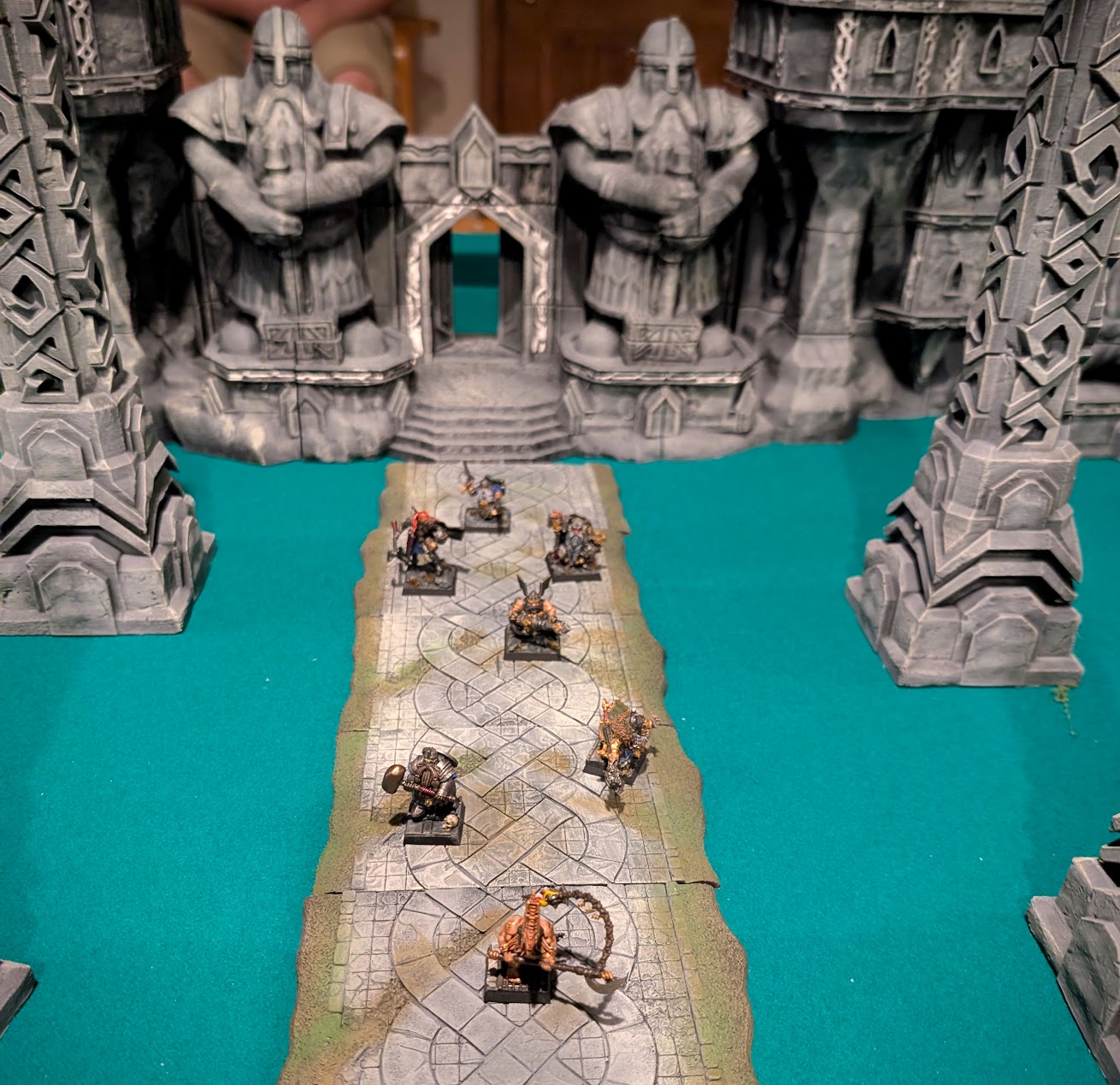 Painted dwarf hero miniatures line a stone road leading to fortress gates flanked by giant dwarf statues.